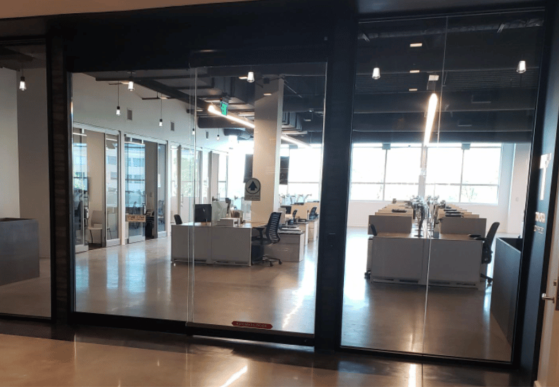 View of a modern open-plan office with desks, chairs, and computers, seen through glass walls; the space is empty and well-lit by large windows, featuring stylish commercial doors that enhance accessibility and design.
