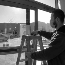 A person wearing a mask stands on a ladder, working near a large glass door inside a building with parked cars visible outside—possibly performing commercial door installation or repair.