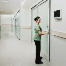 A healthcare worker in green scrubs stands in a hospital corridor, opening a patient room door maintained through regular automatic door maintenance.