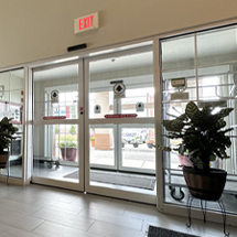 The glass entrance features sliding doors with a clearly marked exit sign above, flanked by potted plants and offering a view of the outside parking area. This elegant setup is supported by reliable automatic door service to ensure seamless operation.