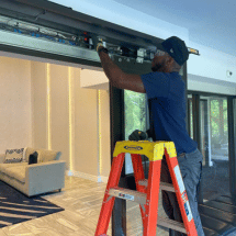Person standing on a ladder performs automatic door repair or inspects a sliding door mechanism in a modern living room with a couch in the background.