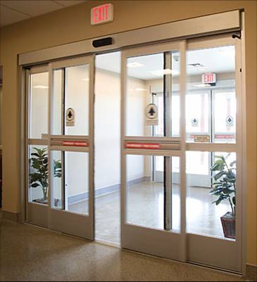 Automatic sliding glass doors at a building entrance, with potted plants on either side and exit signs above, showcasing expert commercial door installation for a welcoming and secure entryway.