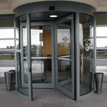 A glass revolving door at the entrance of a building, installed as part of a commercial door installation, with two metal bins on either side and a view of the outside through the glass.