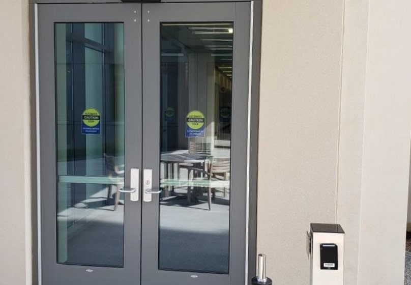 Glass double doors with metal handles and safety labels lead to an interior hallway, highlighting the importance of reliable commercial door repair. A card reader is mounted on the wall beside these secure entryways.